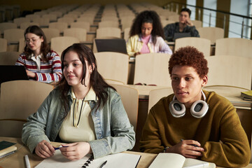 Group of multiethnic university students sitting in lecture hall listening to lesson, two teenagers in foreground taking notes with notebooks and laptops, others studying in background