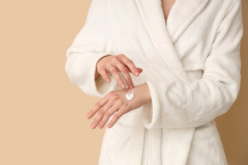 Woman in white bathrobe applying cream onto hands on color background, closeup