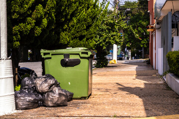 Green dumpster and black trash bags on a sidewalk in a leafy urban street. Illustrates waste management, urban hygiene, and environmental awareness.