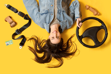 Happy young female driver with license, seat belt and steering wheel on yellow background