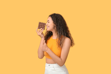 Happy young African-American woman with bar of chocolate showing thumb-up on orange background