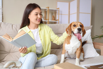 Young woman with cute Beagle dog reading book on sofa in living room