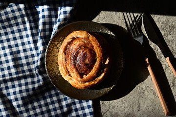 Top view of Danish roll on the plate with sun light, shadows, fork, knife and napkin. Dark, black, pastry, sweet bun, sugar, bread. Grey or gray cement on the background. High angle, above. 