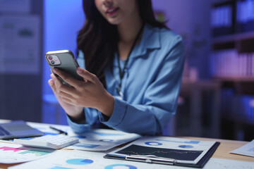 Businesswoman using smartphone and analyzing financial charts at night in office