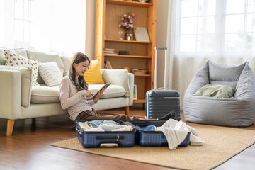 Asian woman traveler checking list on tablet preparing luggage packing clothes in suitcases for...