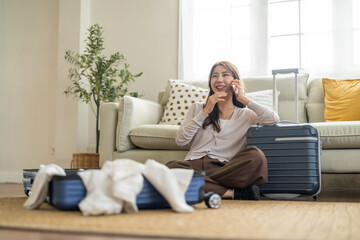 Fototapeta premium Asian woman traveler talking with smartphone and packing clothes in suitcases for travel at home, connected traveler, modern lifestyle, planning holiday travel, content creator and travel blogger