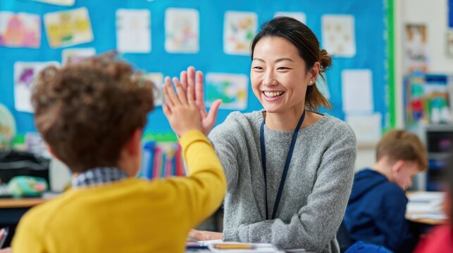 Female Asian teacher high-fiving elementary school student in a bright classroom, colorful posters on the wall, joyful education moment