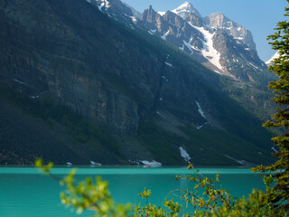Obraz premium Turquoise waters of Lake Louise shimmer beneath towering cliffs and alpine peaks in Banff National Park. A peaceful summer view framed by vibrant greenery and distant snow patches.