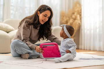 African mother sitting on floor showing tablet to her baby daughter at home, spending time learning...