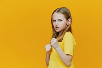 A young girl with long blonde hair, wearing a yellow shirt, shows a fierce expression against a...
