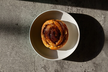 Top view of Danish roll on the plate with sun light and shadows. Dark, black, pastry, sweet bun, sugar, bread. Grey or gray cement on the background. High angle, above. close up.