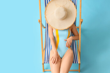 Young woman in swimsuit with hat sitting on deck chair against blue background, top view