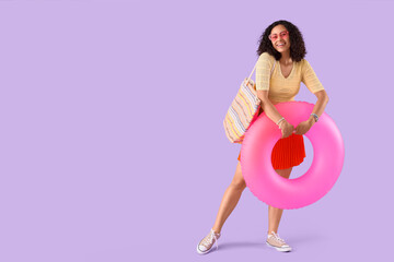 Young African-American woman with pink swim ring and beach bag on lilac background