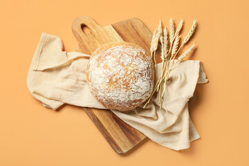 Cutting board with loaf of fresh bread and wheat ears on orange background