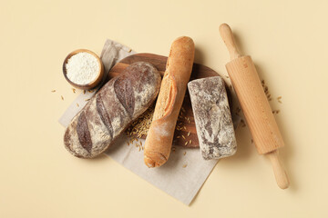Board with loaves of fresh bread, flour and wheat on beige background