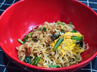 A bright red bowl filled with delicious fried noodles with sliced ​​egg, mustard greens, crispy...