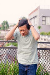 Asian Man Experiencing Frustration or Pain Outdoors Holding Head with Overcast Weather Backdrop