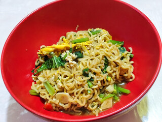 A bright red bowl filled with delicious fried noodles with sliced ​​egg, mustard greens, crispy...