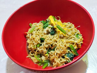A bright red bowl filled with delicious fried noodles with sliced ​​egg, mustard greens, crispy fried onions and sliced ​​garlic, served on a tablecloth.