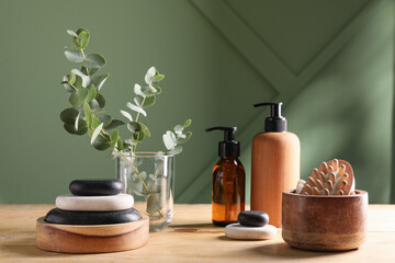 Bottles of cosmetic products, spa stones and bath supplies on wooden table near green wall
