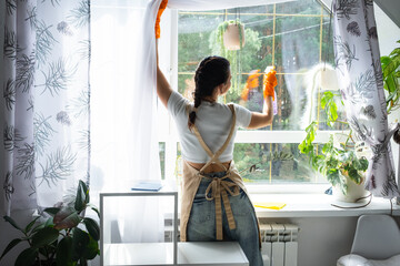 Woman in rubber gloves and apron manually washes window of house with rag cleaner and mop inside interior with home plants on windowsill. Restoring order and cleanliness in spring, cleaning servise