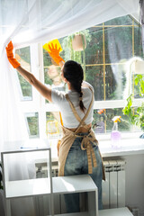 Woman in rubber gloves and apron manually washes window of house with rag cleaner and mop inside interior with home plants on windowsill. Restoring order and cleanliness in spring, cleaning servise