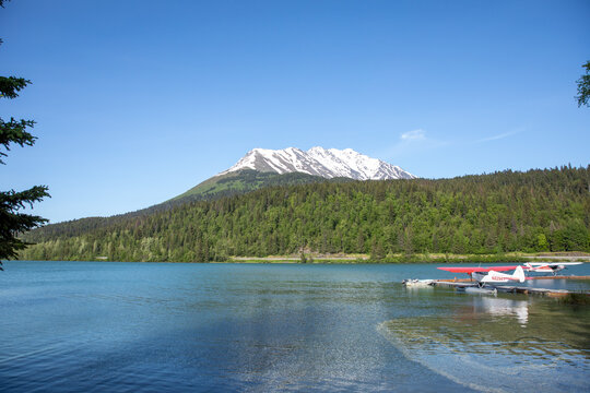 Scenic summer view of small floatplanes docked on a turquoise lake surrounded by dense forest and a snowcapped mountain in Alaska, USA. A classic image of adventure, wilderness, and aviation.