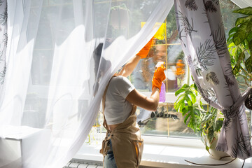 Woman in rubber gloves and apron manually washes window of house with rag cleaner and mop inside interior with home plants on windowsill. Restoring order and cleanliness in spring, cleaning servise