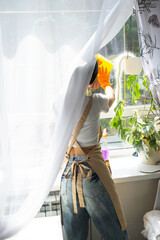 Woman in rubber gloves and apron manually washes window of house with rag cleaner and mop inside interior with home plants on windowsill. Restoring order and cleanliness in spring, cleaning servise