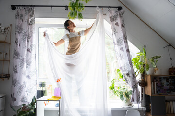 Woman in apron hangs transparent tulle curtains on large attic windows in the house inside the...