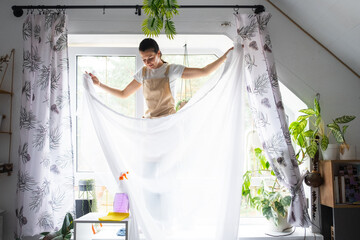Woman in apron hangs transparent tulle curtains on large attic windows in the house inside the interior with potted plants. Spring cleaning, tidying up