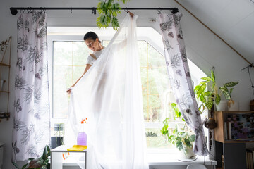 Woman in apron hangs transparent tulle curtains on large attic windows in the house inside the interior with potted plants. Spring cleaning, tidying up