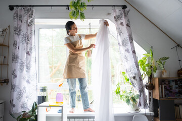 Woman in apron hangs transparent tulle curtains on large attic windows in the house inside the interior with potted plants. Spring cleaning, tidying up