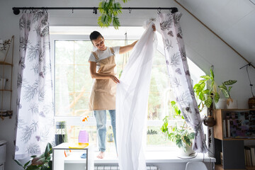 Woman in apron hangs transparent tulle curtains on large attic windows in the house inside the...