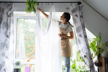 Woman in apron hangs transparent tulle curtains on large attic windows in the house inside the...