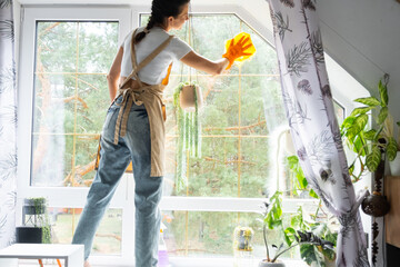 Woman in rubber gloves and apron manually washes attic window of house with rag cleaner and mop...