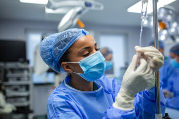 Female anesthesiologist adjusts IV drip in sterile operating room. She is concentrated and careful. Medical staff and equipment appear in the blurred background.