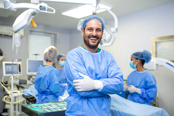 male surgeon stands with arms crossed and a big smile in an operating room while his team prepares...