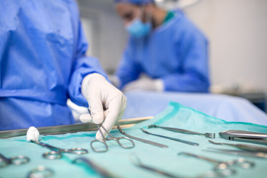 Close-up of a gloved hand selecting a surgical instrument from a sterile tray. In the background, medical staff prepare for the procedure in the operating room.