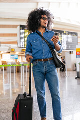 Young man with trolley suitcase using mobile phone and smiling while walking in airport terminal