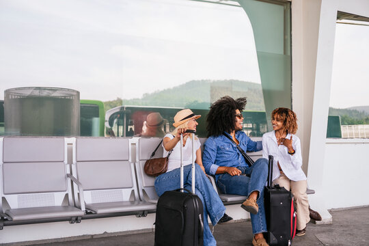 Three multiracial friends are sitting in the waiting area of an airport with their suitcases, chatting and enjoying each other's company