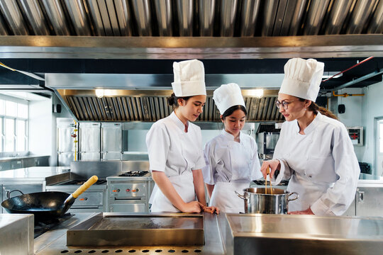Chef instructing two culinary students in white uniforms and tall hats on how to cook in a commercial kitchen, standing near a pot on the stove during a hands-on professional cooking class.