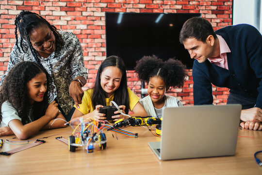 Diverse group of students learning robotics with teachers in a classroom. Smiling girls use controller and laptop to operate a robotic arm. Fun STEM activity promotes coding, teamwork, and curiosity.