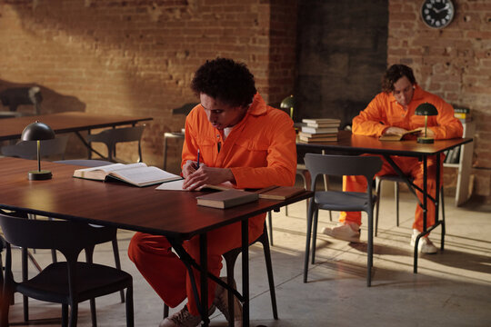 Young adult man sitting at table writing in notebook while young adult Caucasian man reading book in background, both wearing orange prison uniforms in prison library setting