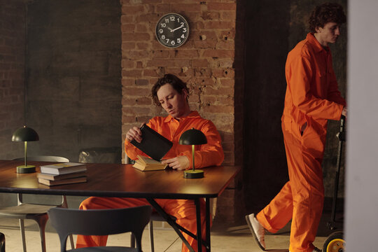 Two men wearing orange jumpsuits spending time in prison library, one sitting at table reading book while other walking past, brick wall and clock in background