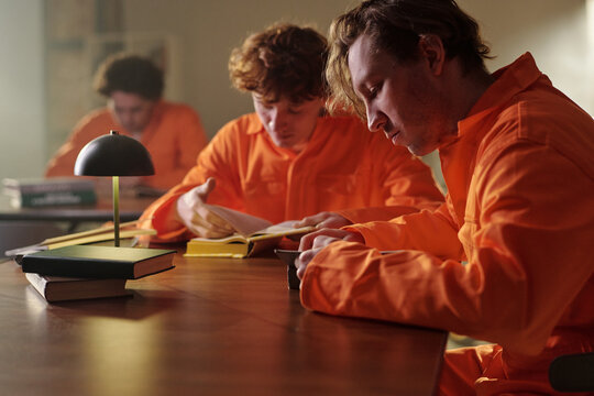 Three young adult men wearing orange uniforms sitting at table reading books in prison library, focused on studying and learning, books and desk lamp visible in foreground