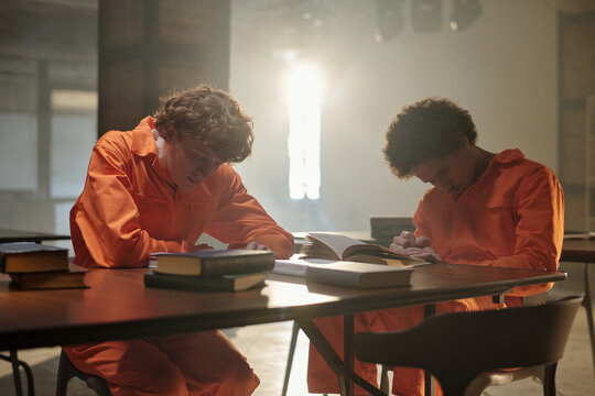 Two men sitting at table in prison library, reading books and writing notes, focused on studying, surrounded by stacks of books on desk