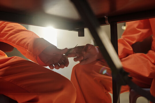 Two young adult men in orange prison uniforms sitting under table exchanging small object with hands visible, scene taking place in prison library setting with focus on secretive action