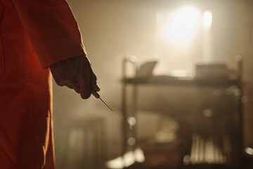 Man holding sharp object in hand standing in prison library with books and metal cart visible in background, focus on hand and weapon, face not visible