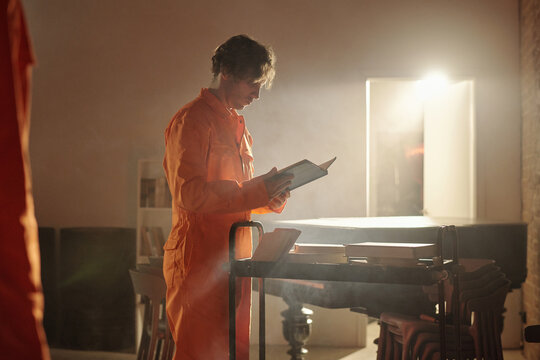 Young adult Caucasian man standing in prison library reading book while holding cart with stacked books, focused on pages, wearing orange jumpsuit, illuminated by bright light